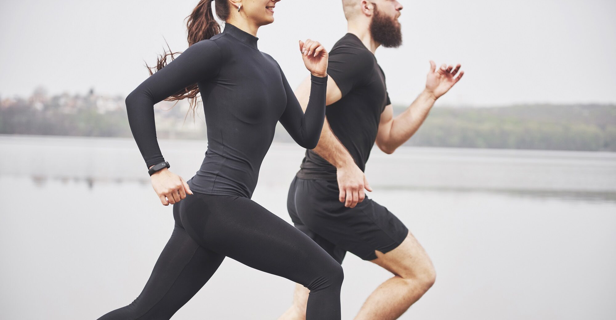 Couple jogging and running outdoors in park near the water. Young bearded man and woman exercising together in morning.